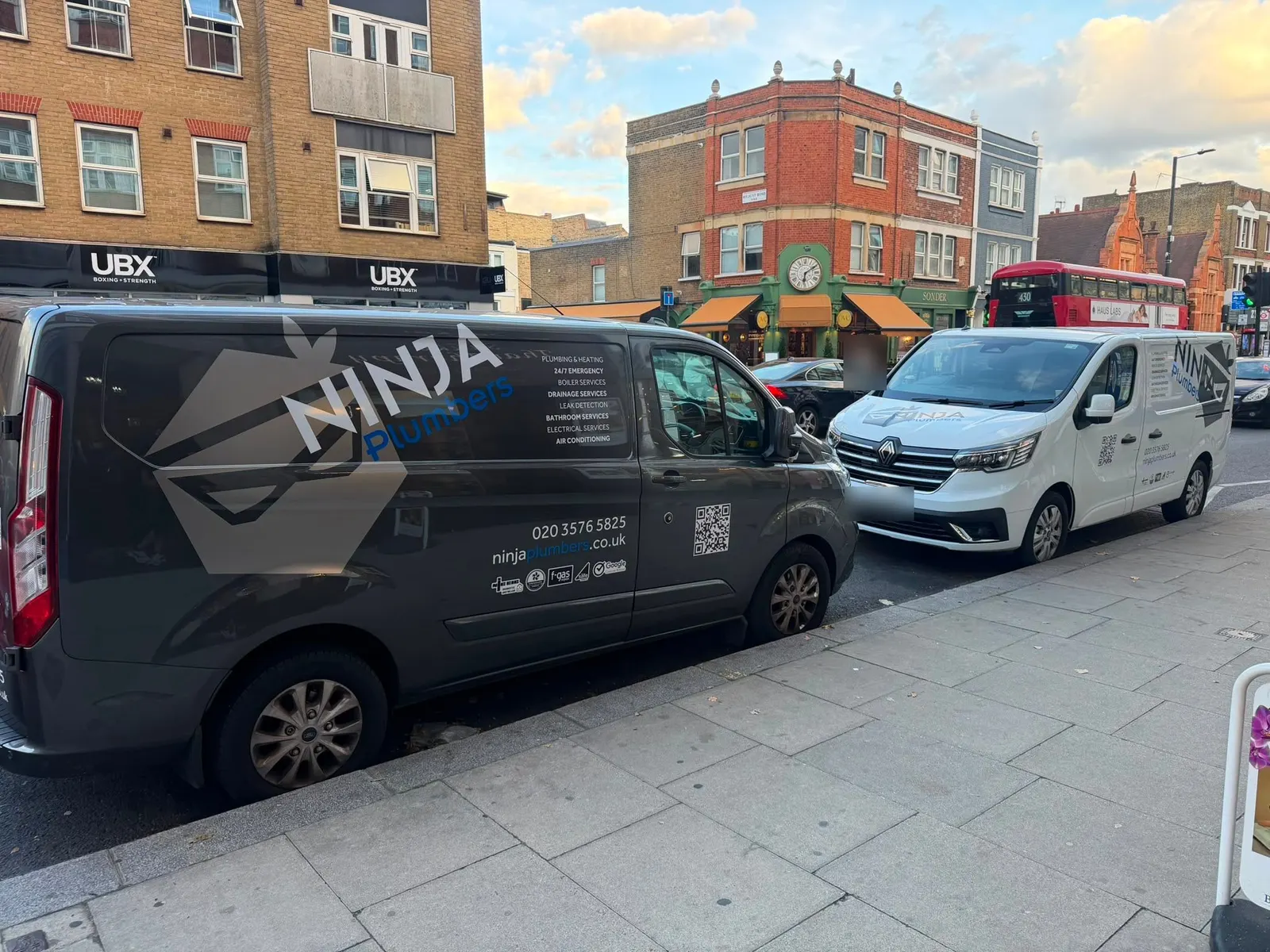 A dark grey van and a white van in Ninja Plumbers livery, showing the phone number and website, parked on a London high street